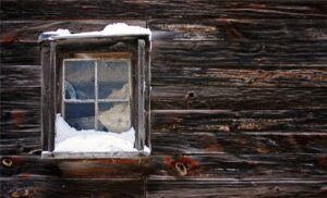 Small window in an old wooden cabin wall with snow on the sill